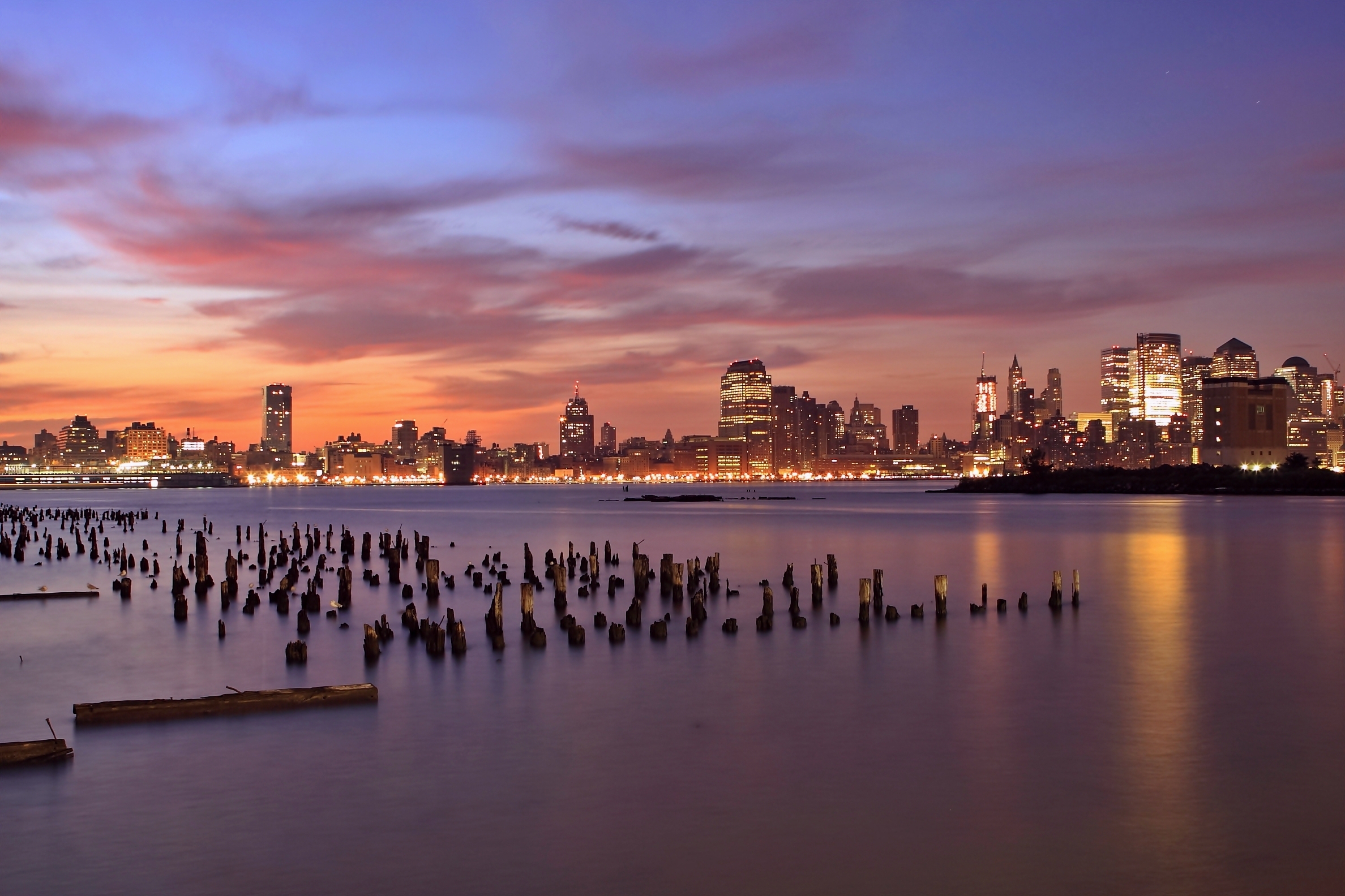 cities, rivers, sunset, sky, lilac, clouds, usa, orange, lights, wooden, skyscrapers, backlight, illumination, evening, united states, pillars, jersey city, state of new jersey, new jersey state, hudson, columns cell phone wallpapers